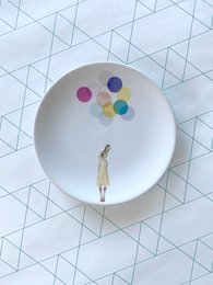 A white ceramic plate with a whimsical illustration of a person holding colorful balloons.