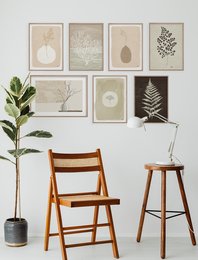 A set of minimalist, nature-inspired art prints featuring various botanical elements such as leaves, branches, and organic shapes in neutral tones. The prints are displayed in a gallery-style arrangement on the wall, complemented by a potted plant and simple wooden furniture.