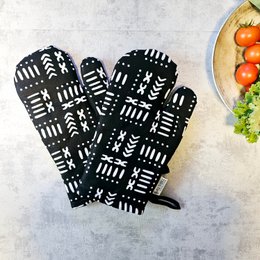 A pair of black and white oven mitts with a bold, geometric print inspired by traditional African Mudcloth textiles. The oven mitts feature a variety of shapes and patterns, including arrows, lines, and abstract designs, creating a striking and visually interesting appearance. These oven mitts would be a stylish and functional addition to any kitchen.
